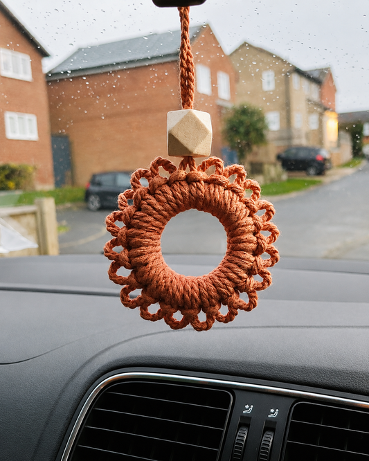 Handmade macrame daisy car charm hanging from a rear view mirror with wooden diffuser bead, adding boho style and natural charm to a car interior.