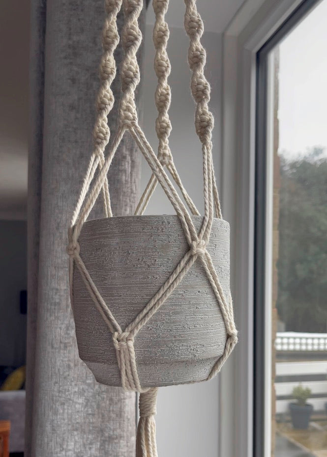 Macrame plant hanger with a gray pot hanging from it indoors.