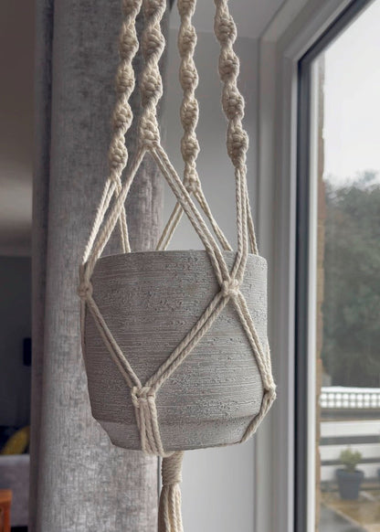 Macrame plant hanger with a gray pot hanging from it indoors.