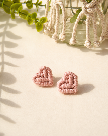 Blush pink handmade macramé heart stud earrings on a neutral fabric background.