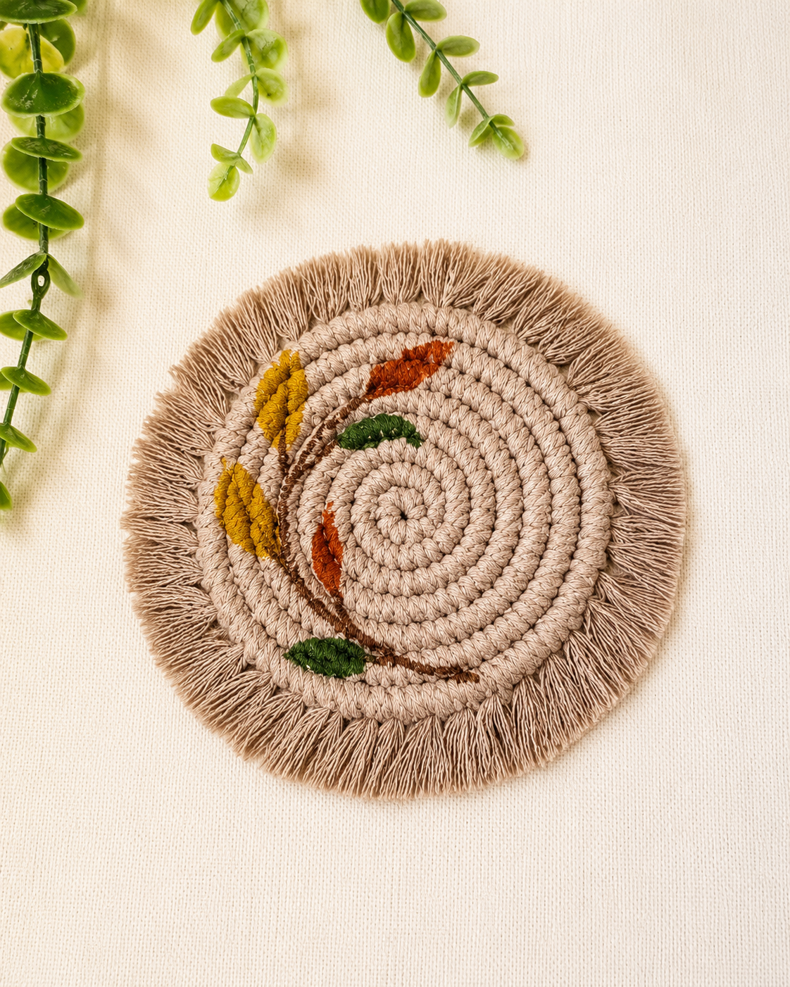 Boho floral macrame coaster handmade from cotton cord with embroidered leaf design and fringe in beige and earthy tones.