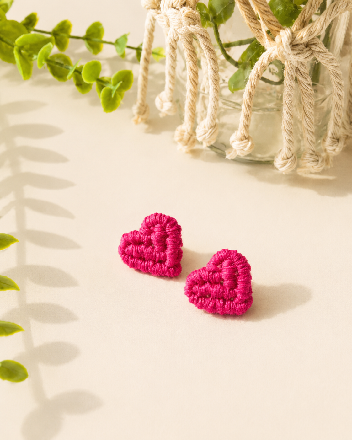 Fuchsia pink handmade macramé heart stud earrings on a neutral fabric background.