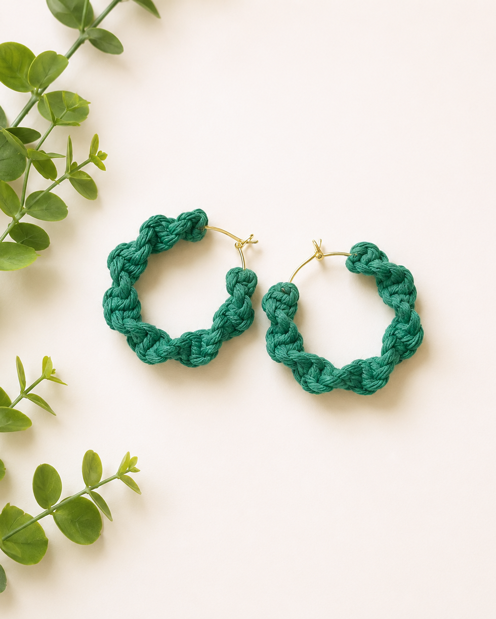 Handmade green macrame hoop earrings on an off-white background with green foliage. Lightweight boho cotton earrings for women with a modern natural style.