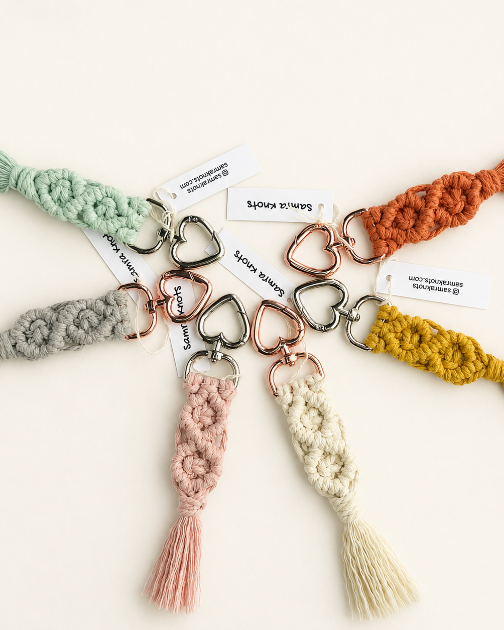 Flat lay of seven handmade macramé diamond keychains arranged in a circle, each in a different colour with heart-shaped metal clasps on a soft neutral background
