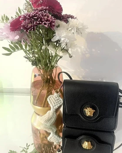 Black handbag with gold emblem next to a vase of flowers on a reflective surface