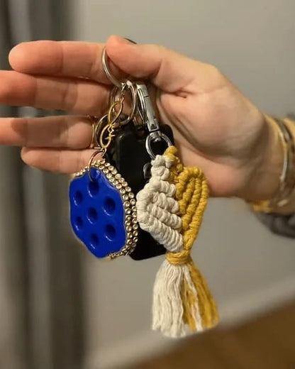 Hand holding a keychain with a blue and black object and a white tassel against a neutral background