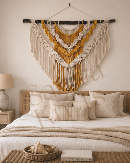 Large off-white and mustard macrame wall hanging styled above a bed in a light bedroom.