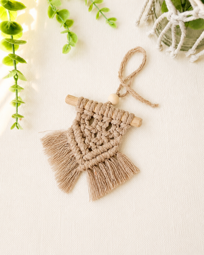 Handmade light brown mini macrame wall hanging with wooden dowel and fringe displayed on an off-white background.