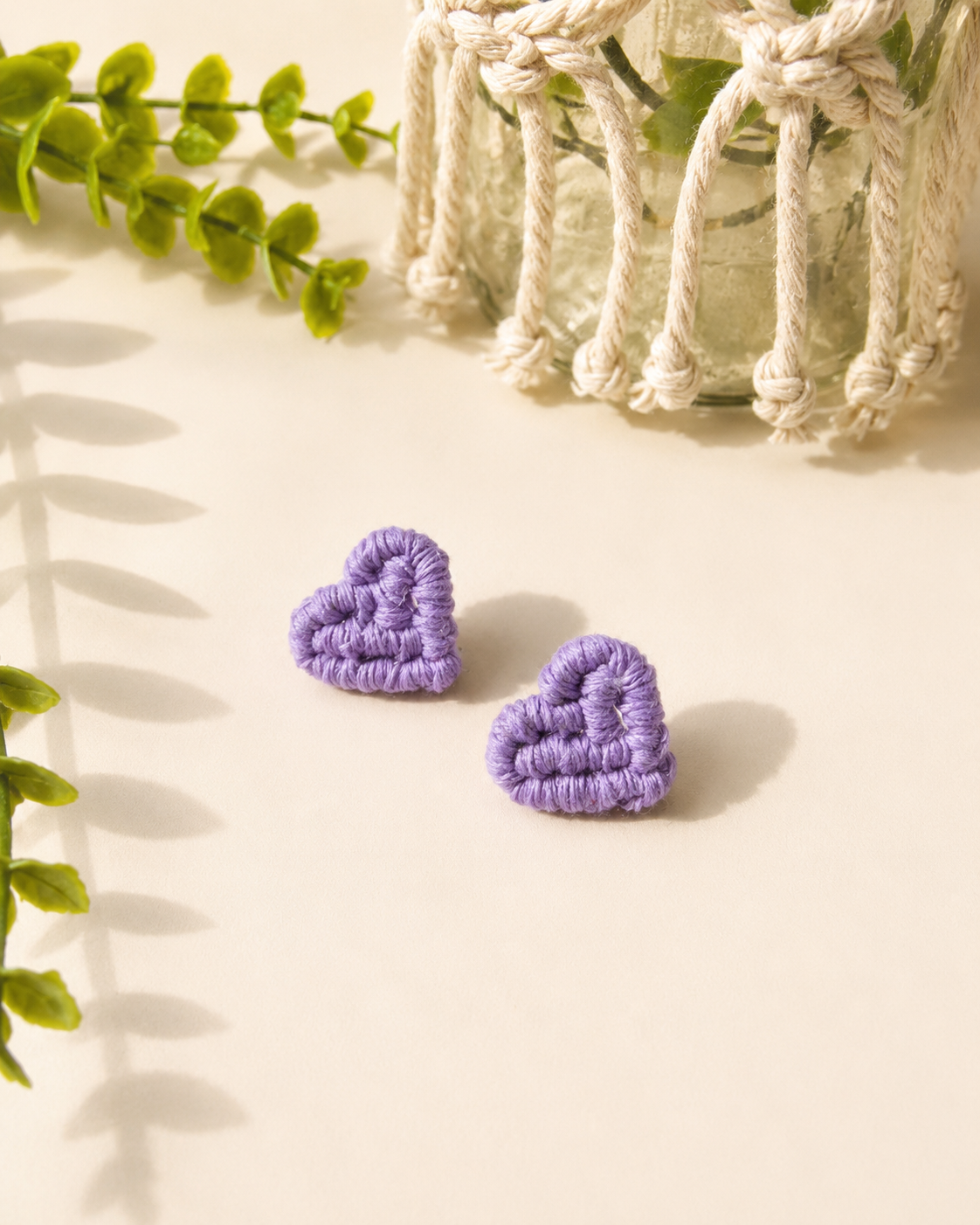Lilac handmade macramé heart stud earrings on a neutral fabric background.