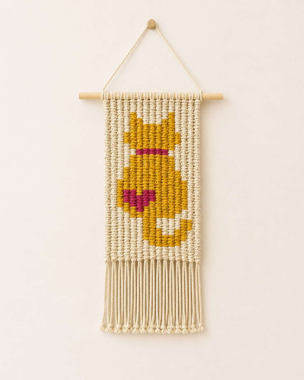 Handmade macrame cat wall hanging with mustard yellow cat design and pink heart, woven from cotton cord and displayed on a wooden dowel.