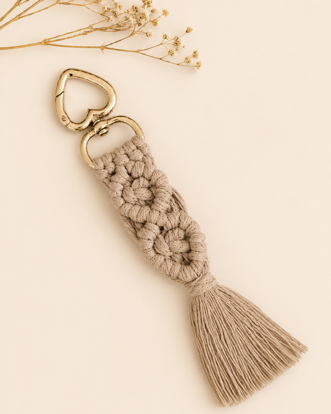 An earthy brown macrame keychain with a gold-tone heart-shaped clasp and tassel, styled on a light beige background with dried flowers in the top corner.