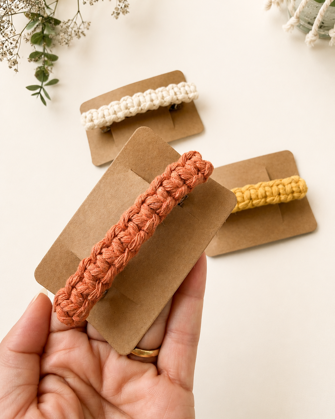 Handmade terracotta macrame hair clip displayed on a kraft backing card, held in hand with ivory and mustard macrame hair clips in the background.