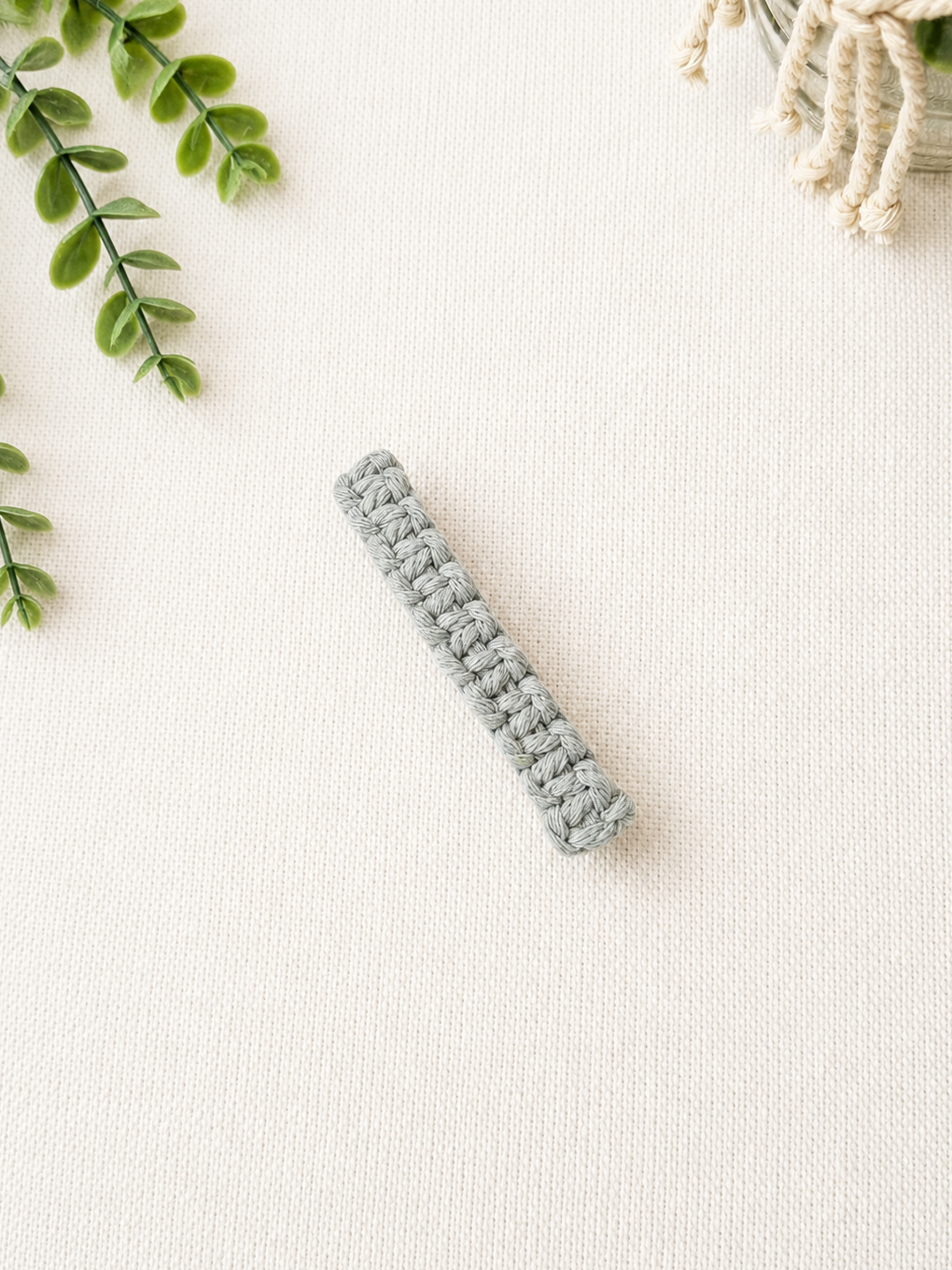 Handmade sage green macrame hair clip on an off-white background with greenery and a decorative macrame planter.