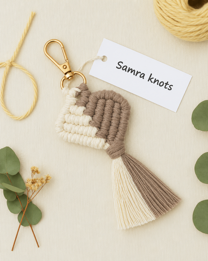 A handmade macrame keychain with a gold clasp, featuring cream and taupe woven cords and a tassel, displayed on a textured cream background with decorative eucalyptus leaves, a spool of yellow cord, and a paper tag reading 'Samra knots'.