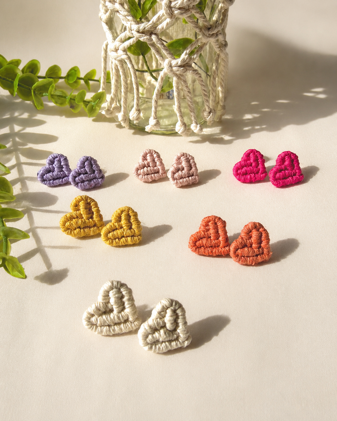 Handmade macramé heart stud earrings in multiple colours displayed on a neutral fabric background with boho styling.