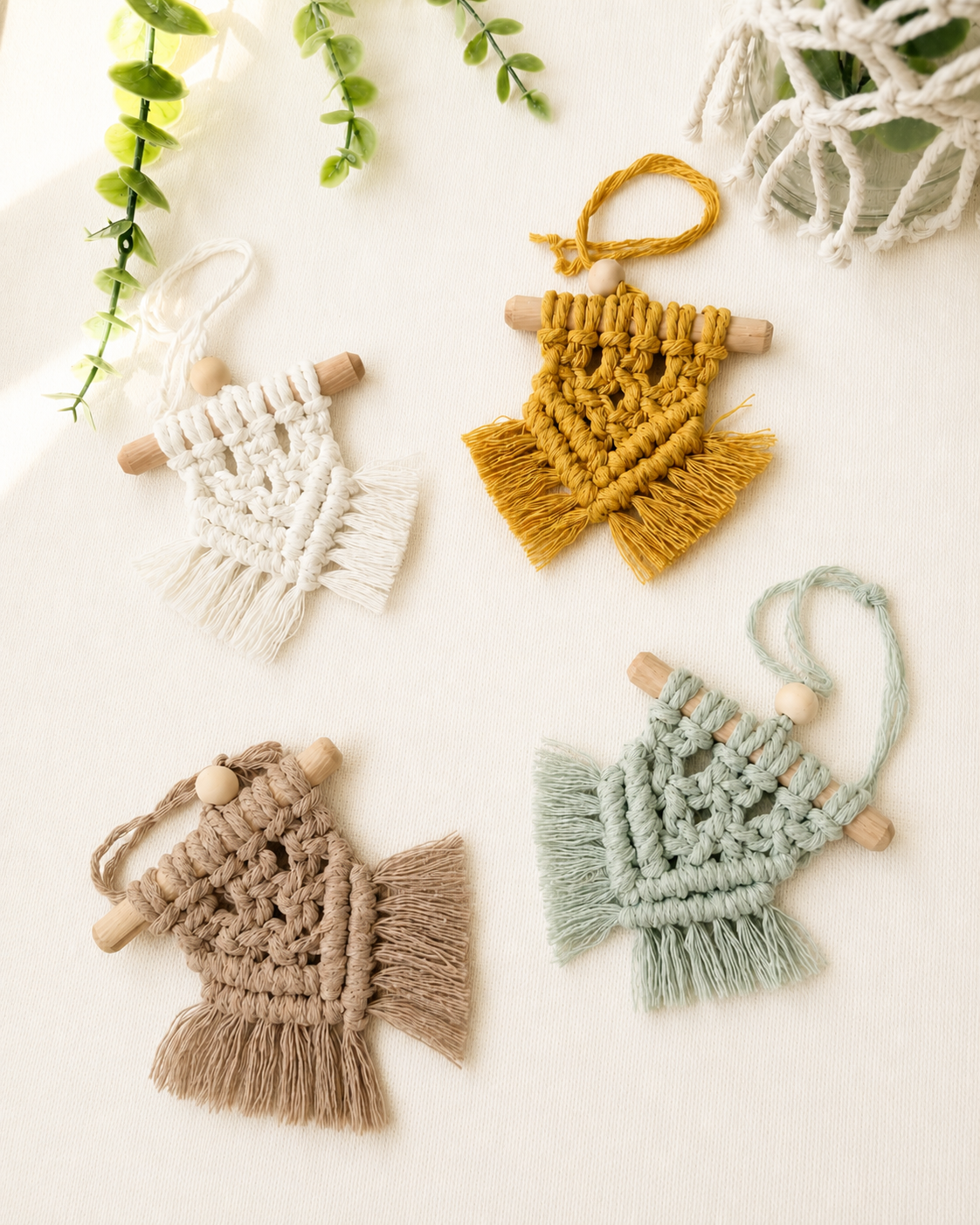 Set of four handmade mini macrame wall hangings in off white, mustard yellow, mint green and light brown displayed on an off-white background with greenery.