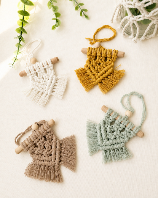 Set of four handmade mini macrame wall hangings in off white, mustard yellow, mint green and light brown displayed on an off-white background with greenery.