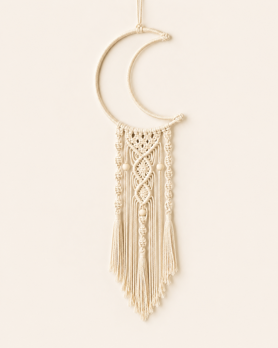 Handmade macrame crescent moon wall hanging in natural cotton with decorative knots, wooden beads and tassel fringe on a warm off-white background.