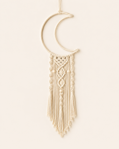 Handmade macrame crescent moon wall hanging in natural cotton with decorative knots, wooden beads and tassel fringe on a warm off-white background.