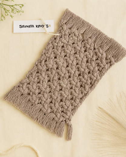 A chunky knit cable scarf in taupe brown with fringed ends, laid flat on a light cream background with a small tag reading 'samra kno' visible.