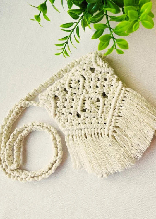 Handmade off-white macramé crossbody bag with fringe and intricate knot design, styled with green leaves – Samra Knots.