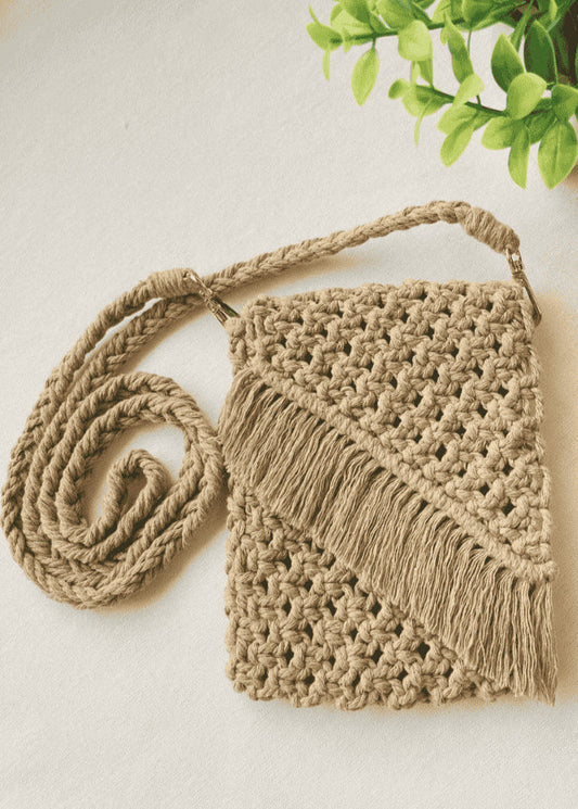 Full flat-lay of a light brown macramé crossbody bag with braided strap and fringe, displayed with green plant decor on neutral background.