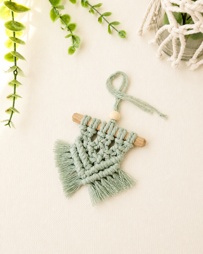 Handmade mint green mini macrame wall hanging with wooden dowel and fringe displayed on an off-white background.