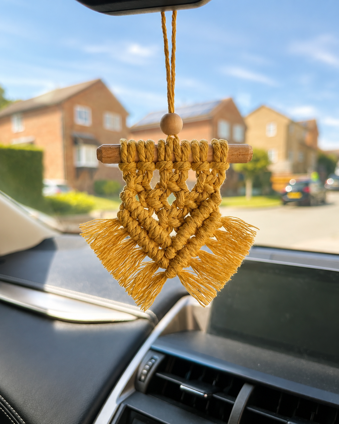 Handmade mustard macramé car charm hanging from a rear-view mirror inside a car with a softly blurred background.