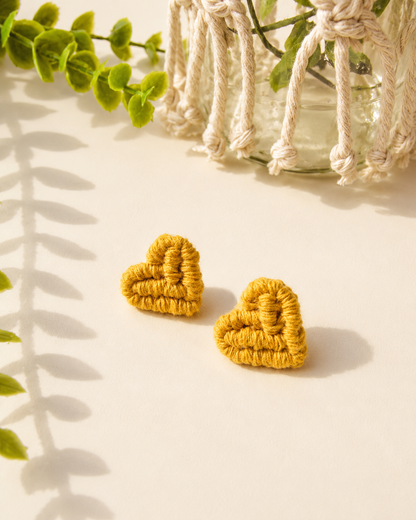 Mustard yellow handmade macramé heart stud earrings on a neutral fabric background.