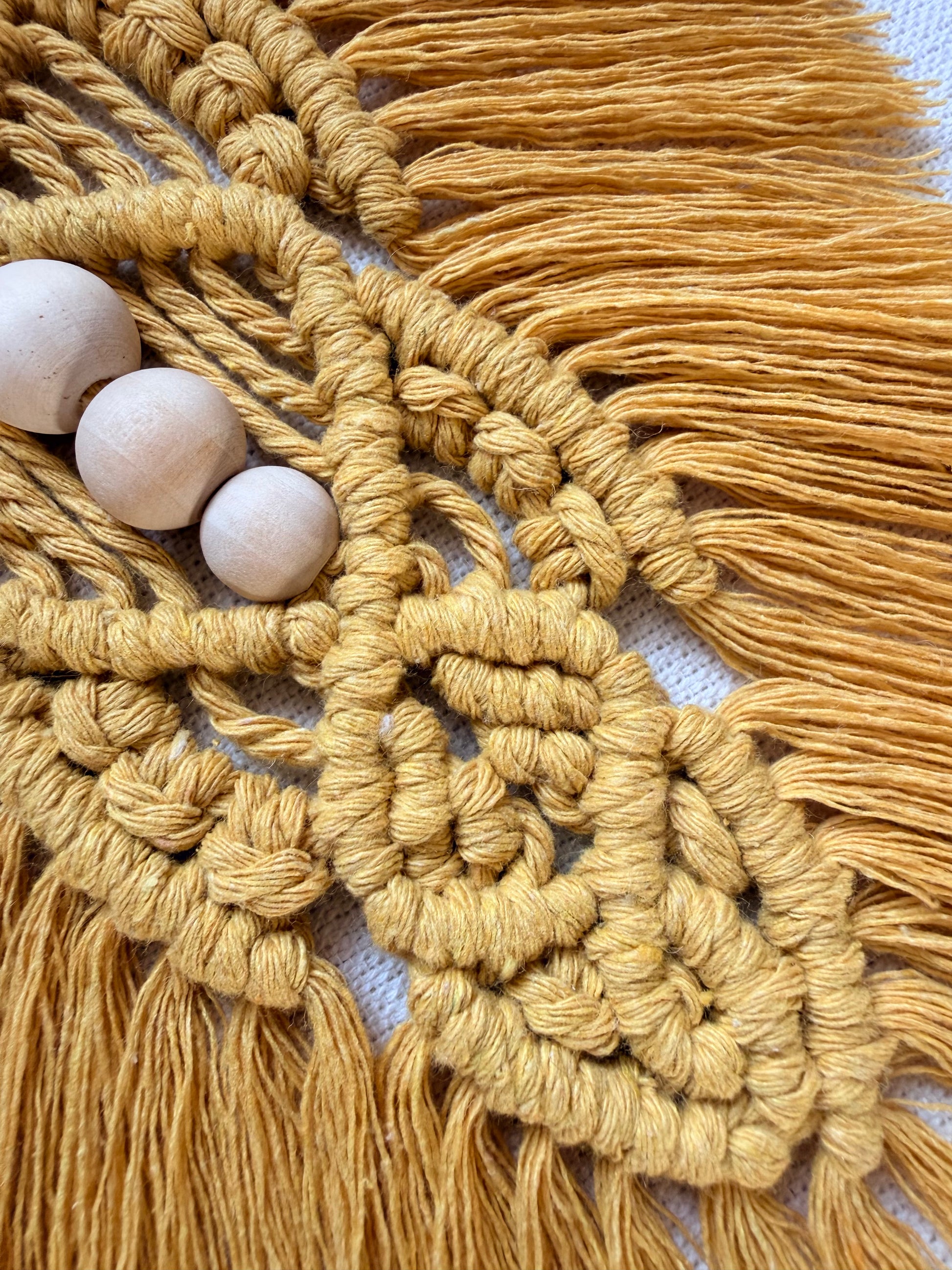 Close-up of a handmade mustard macrame wall hanging featuring intricate knotwork, natural wooden beads and soft cotton fringe.