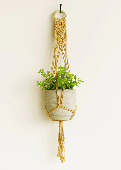 Mustard yellow macrame plant hanger handmade with cotton cord and wooden ring holding a small plant against a neutral background.