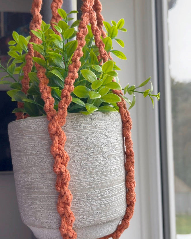 Close-up of a terracotta macrame plant hanger featuring handmade half hitch knots and a textured grey planter with green foliage.